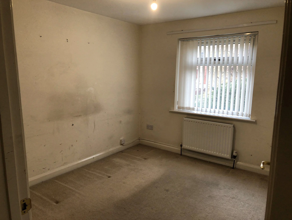 An empty, unfurnished bedroom with bare walls, worn carpet and a window with vertical blinds